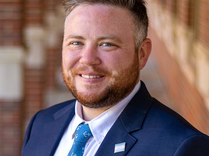 A traditional business headshot of Aiden in a navy-blue suite with a trans pride flag pin on their lapel