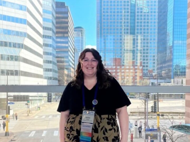Brittany stands, hands to her side and wearing a conference lanyard, in front of a picture window, apparently on a catwalk above a street.
