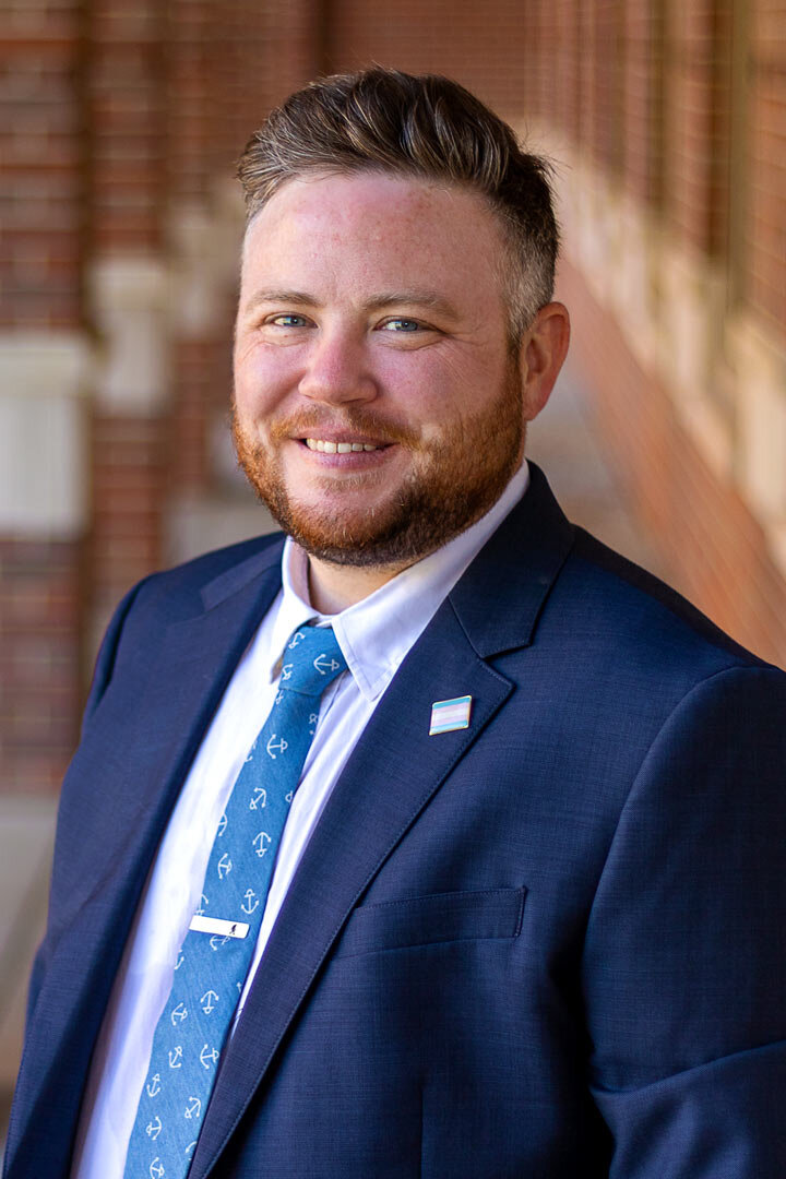 A traditional business headshot of Aiden in a navy-blue suite with a trans pride flag pin on their lapel