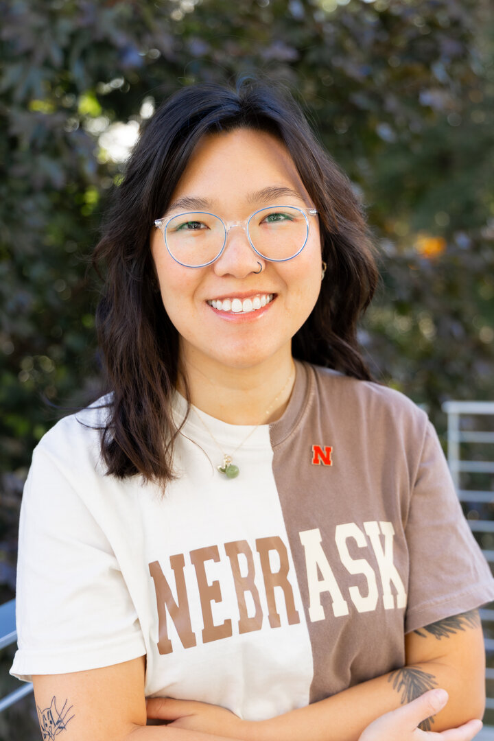 Justine stands, loosley crossed armed and smiling, in a two-tone Nebraska t-shirt (cream on her right and soft brown on her left) and a Nebraska "N" pin near the neck of the shirt on the left side.